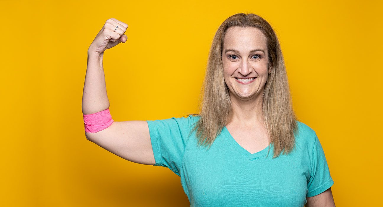 胆管癌症护理人和献血者梅利莎·富奎·贝克（Melissa Fuquay Baker）站在黄色的背景前，一边握住手臂，在捐献血后展示粉红色的绷带。
