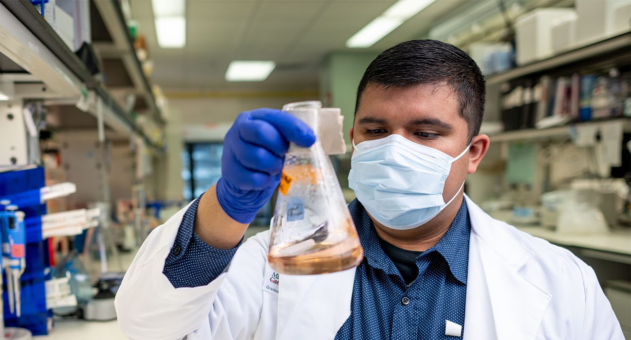 研究生Rolando Vedia戴着白色外套，医疗级面罩和蓝色手套，在实验室里握住烧杯。