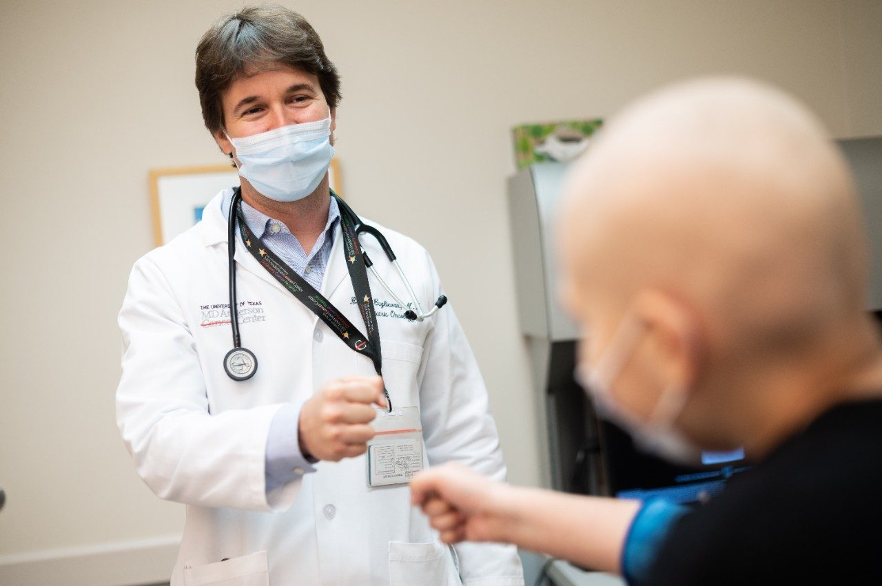 医学博士布兰科·库格里埃文(Branko Cuglievan)身穿白大褂，戴着医用口罩，在诊所里与一名儿童白血病患者碰拳