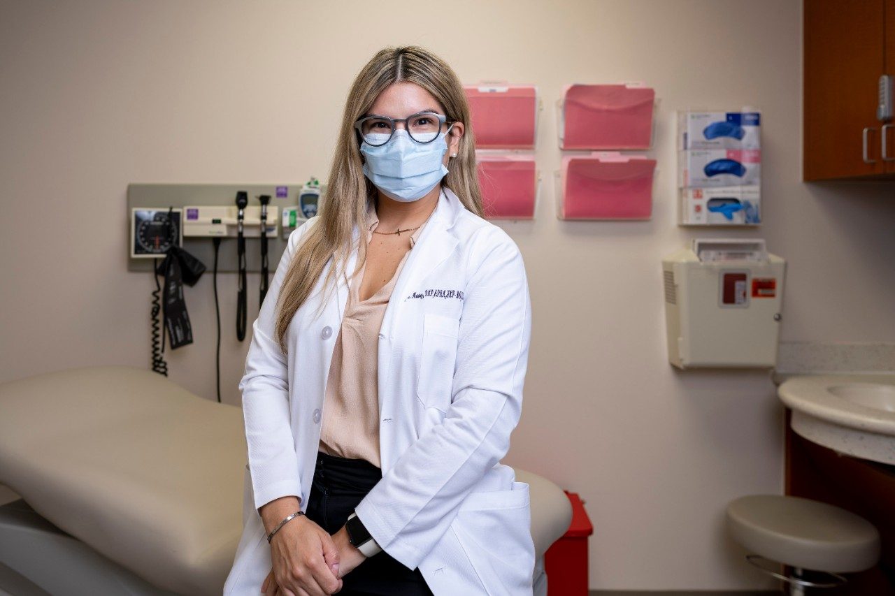 拉丁裔护士阿什莉·马丁内斯(Ashley Martinez)坐在一间诊室里，双手叠在一起，身后挂着文件夹。