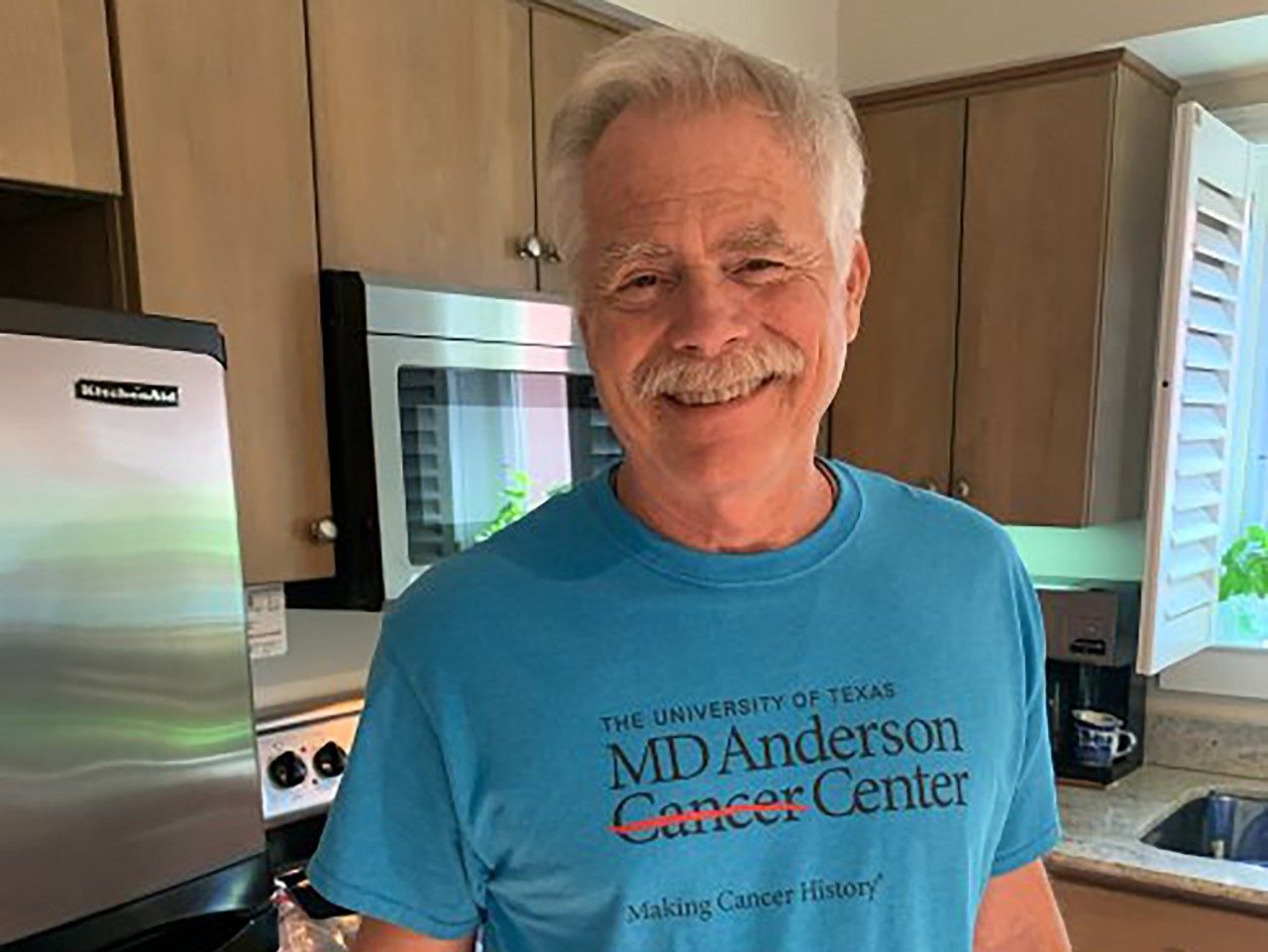 帕特里克·米德（Patrick Mead）在他的厨房里微笑着，一件蓝色的T恤和MD Anderson徽标上的蓝色T恤。他是一个年长的白人。