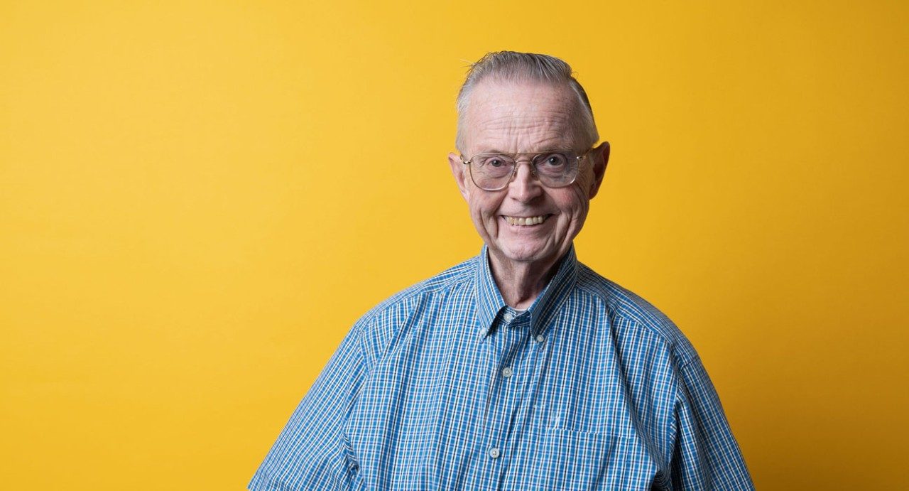 照顾者和捐助者威廉·布雷迪（William Brady）站在黄色背景前，微笑着。他是70多岁的男人，戴着眼镜和蓝色的纽扣衬衫。