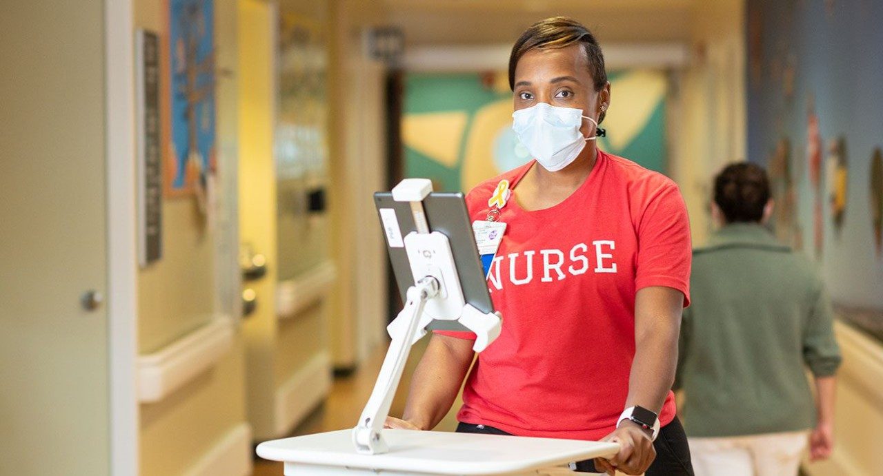医院护士Tiambe Kuykendall戴着医学级的口罩和红色T恤，站在MD Anderson的走廊上的iPad前面，上面写着“护士”。