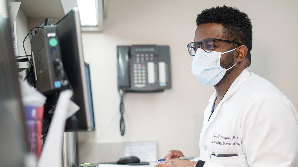 M.D. Uzondu Osuagwu看着一台计算机监视器，穿着他的白色外套和医疗级面罩