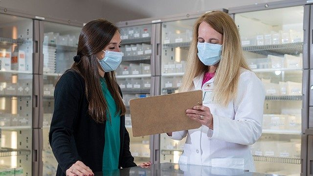 药房经理Stefani Gautreaux（右）在与药房的药房居民劳伦·戈德堡（Lauren Goldberg）交谈时，在药房里举起了剪贴板。这两个女人都有长发，并且戴着医学级面具。