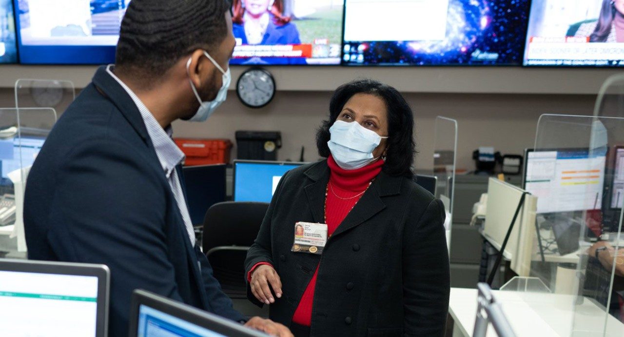 德维娜·帕特尔（Devina Patel）与一位男同事咨询；显示天气预报的电视在后台。两位员工都戴着医学级的口罩。