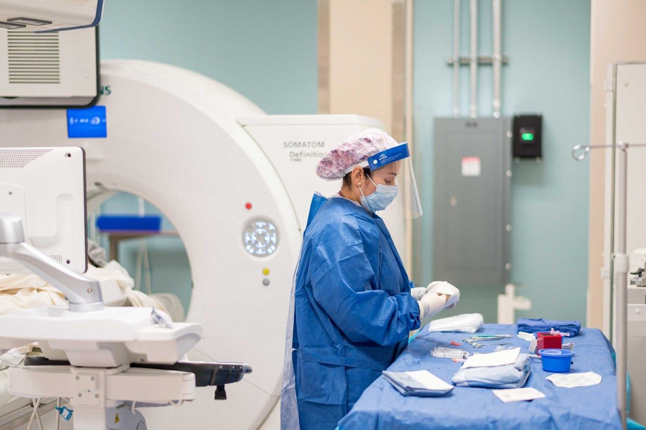 一个人穿着完整的实习医生风云和PPE站在一张桌子前面准备餐具为介入放射学过程。她身后是一个扫描机器。