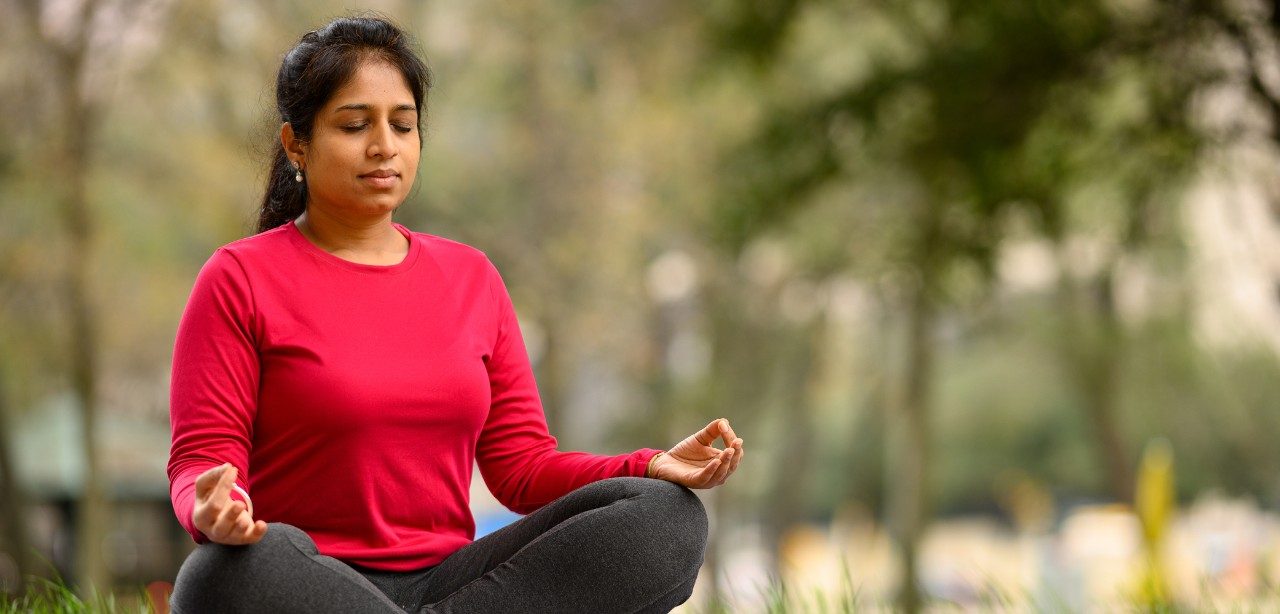 Smitha Mallaiah, MD Anderson Senior Mind/Body Intervention Specialist, is shown meditating outdoors with trees behind..