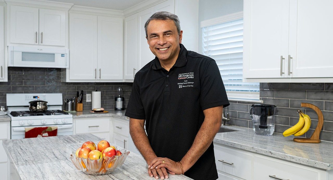 罗纳德·亚伯拉罕（Ronald Abraham）站在他新近翻新的厨房中。他的手放在一个白岛上，而一碗红苹果则坐在他旁边。他在右上角微笑着，穿着带有MD Anderson徽标的黑色领衬衫。