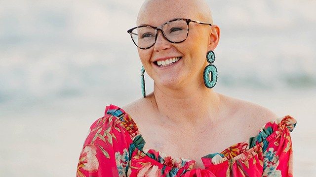 Stage IV Hodgkin lymphoma survivor Mallory Parrish, wearing glasses and a red floral top and big turquoise earrings, smiles while looking off in the distance.