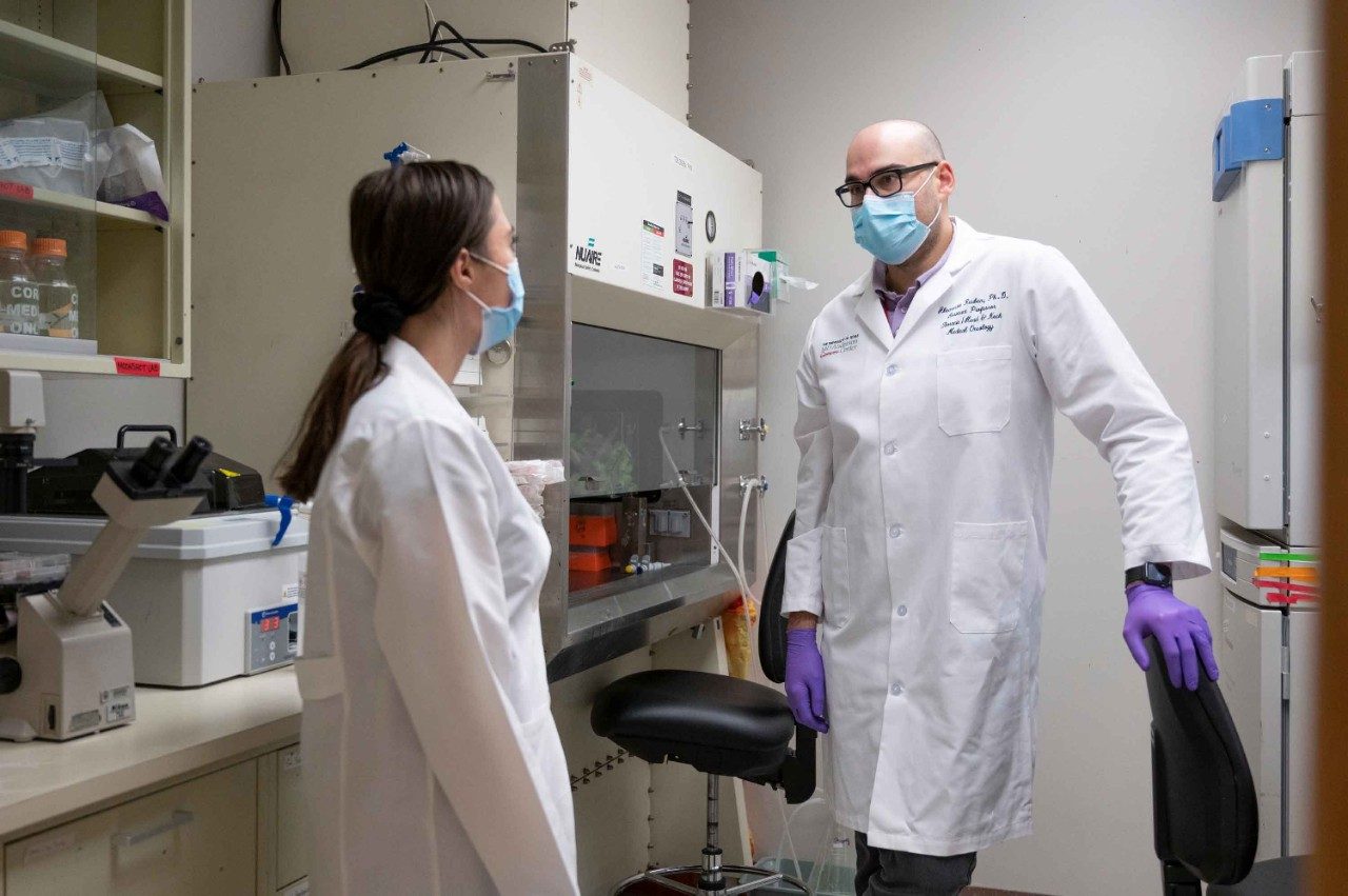 亚历山大·鲁宾博士说话带有女性实验室在实验室成员。两者都是穿着白大褂,紫色乳胶手套和口罩。