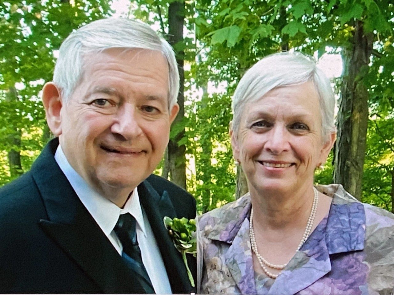 鲍勃Wagar,医学博士,和他的妻子罗宾站在绿树前而穿西装和一个紫色和粉色花上衣和珍珠,分别。他们是一位年长的白人夫妇与灰色的头发。