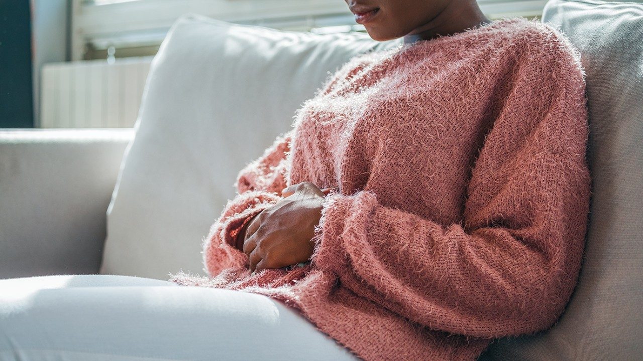 黑人妇女在一个模糊的粉红色毛衣和白色修身长裤抱着她胃疼在白色的沙发上
